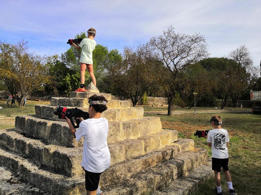 Enfants jouant au Laser Game autour d’une pyramide en pierre, se cachant et visant avec leurs fusils laser.